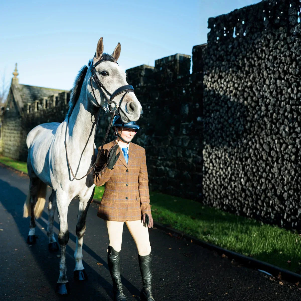 Dapple grey horse wearing an EcoRider leather bridle, presented in-hand by an equestrian handler in a classic tweed jacket.
