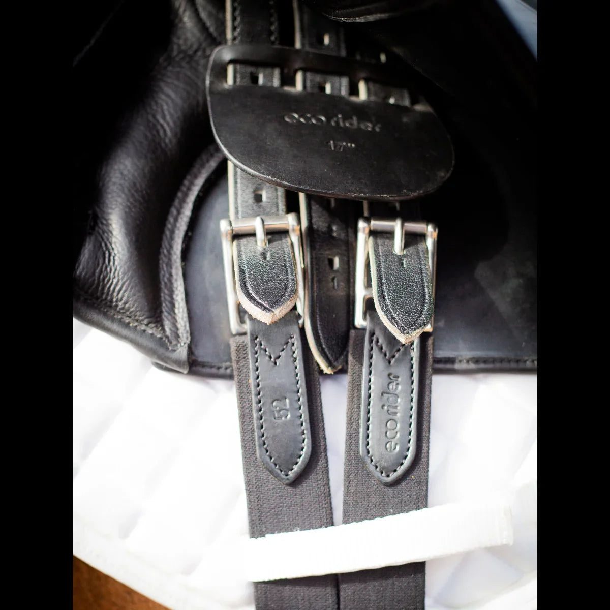 Close-up of the black EcoRider girth buckled to the saddle billets, showing the size 52 stamp and debossed branding.