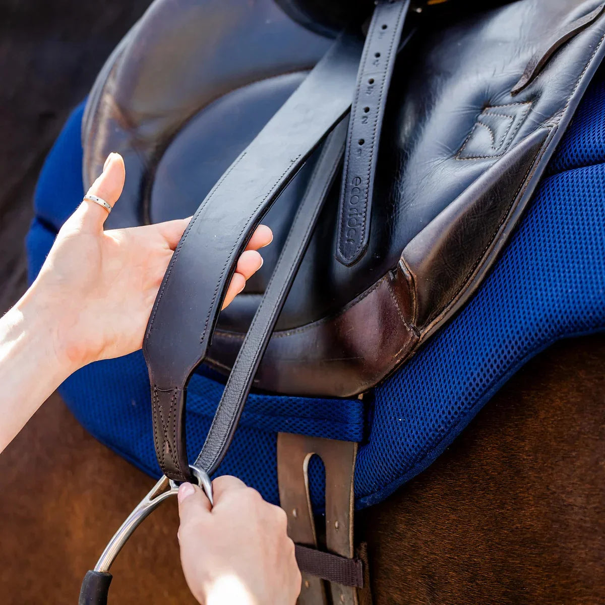 Close-up of a handler adjusting the EcoRider Balance Stirrup Leathers on a saddle, showing the wide-body leather design.